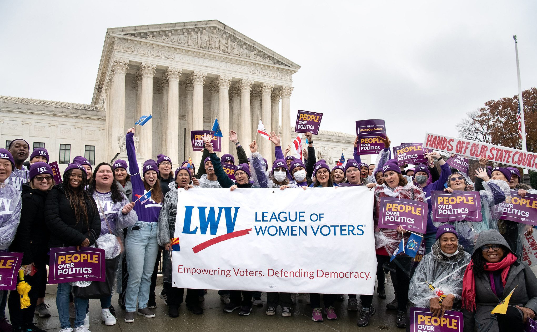 A diverse group of people hold a large "League of Women Voters" banner in front of a courthouse, with signs advocating for democracy and fair politics.