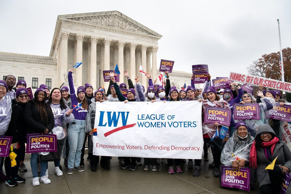 Group of people holding a "League of Women Voters" banner in front of a large building. Participants hold signs with messages about politics and democracy.