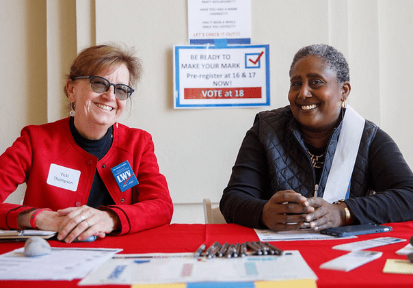 Two women seated at a red table smiling, with registration forms and pens in front. A poster behind them encourages pre-registration for voting at ages 16 and 17.