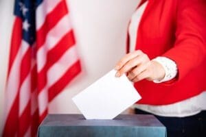 A person in a red jacket casts a ballot into a voting box, with an American flag visible in the background.