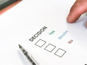 A hand points to a checklist with the word “DECISION” and three options: Yes, Maybe, and No, next to empty checkboxes; a pen lies nearby.