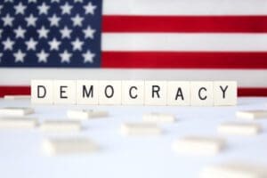 Scrabble tiles spell "DEMOCRACY" in front of a blurred American flag background.