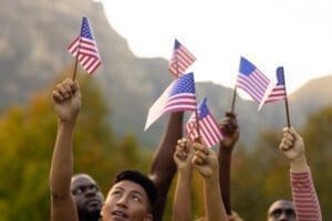 A group of people holding small American flags raised in the air outdoors, with trees and mountains in the background.