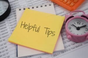 A yellow sticky note with "Helpful Tips" written on it sits on a notepad, next to a pink alarm clock and an orange calculator, over documents.