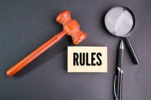 A wooden gavel, a magnifying glass, and a pen are placed around a card that reads "RULES" on a dark surface.