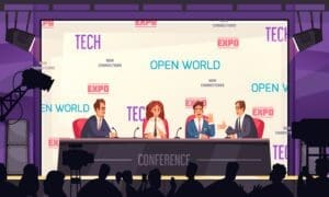 Four people sit at a conference panel table on stage, speaking to an audience. The background displays words like "TECH," "EXPO," and "OPEN WORLD.