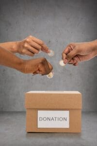 Three hands are shown dropping coins into a cardboard box labeled "DONATION" against a plain grey background.