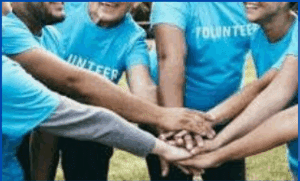 A group of people wearing blue "volunteer" shirts stand in a circle with their hands stacked together in the center.