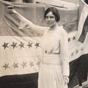 A woman in a white dress raises a glass in front of a large flag featuring stars. Another person holds the flag in the background.