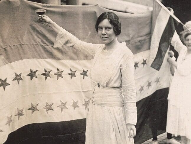 A woman in a white dress raises a glass in front of a large flag featuring stars. Another person holds the flag in the background.