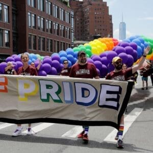 People march in a Pride parade, holding a banner and walking under a large rainbow balloon arch on a city street.