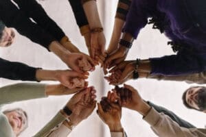 A group of people stand in a circle with their hands extended toward the center, fingers interlocked.
